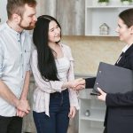A young couple shakes hands with a real estate agent during a home tour or negotiation in a modern setting.